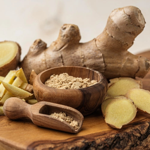 Fresh ginger root, sliced ginger, and ginger powder arranged on a wooden cutting board with a small wooden bowl and scoop.