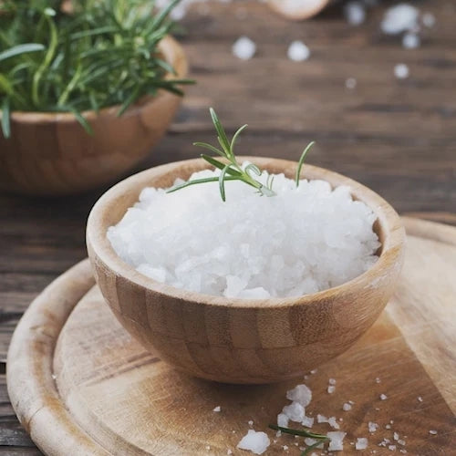 Coarse white sea salt crystals in a small round wooden bowl, garnished with a sprig of rosemary, set on a wooden board with a blurred background of rosemary and more salt.